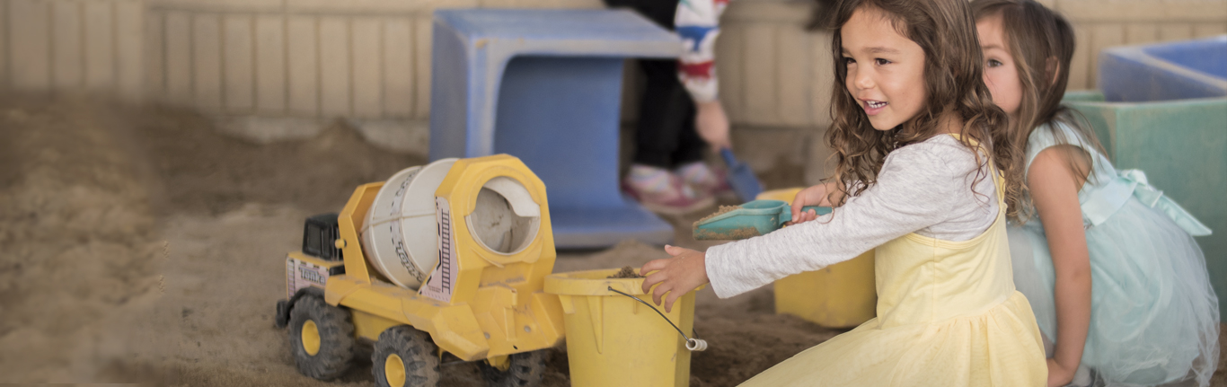 在儿童早期教育项目中，小女孩们穿着裙子在沙盒里玩卡车。