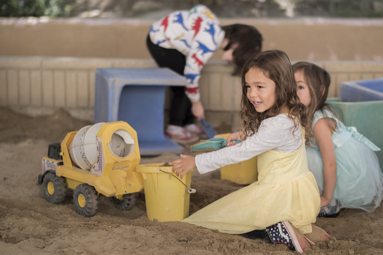 在儿童早期教育项目中，小女孩们穿着裙子在沙盒里玩卡车。图片©Robin Spurs